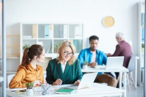 Duas empresárias sentadas à mesa e discutindo a apresentação on-line em equipe com outras pessoas trabalhando em segundo plano. Parecem felizes e seguros graças à nossa lista de produtos para limpeza em escritórios.