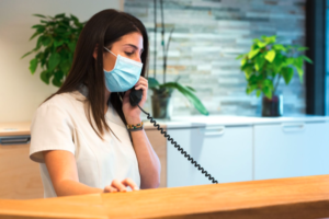 mulher de máscara conversando no telefone em uma mesa de madeira, recepcionista na recepção com uma parede branca ao fundo, forma de prevenir As condições de higiene e cuidado no local de trabalho podem ajudar a prevenir doenças respiratórias durante o inverno. Mas por que a lavagem das mãos e a etiqueta respiratória são tão importantes para manter a saúde e o bem-estar?